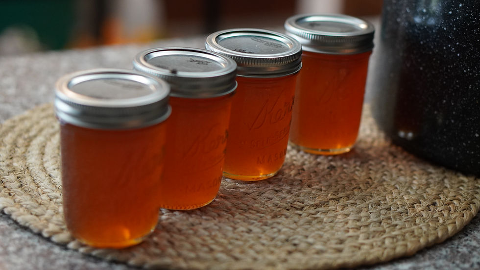 Four mason jars filled with amber liquid sit on a woven mat, next to a dark pot. The scene is cozy, with soft lighting.