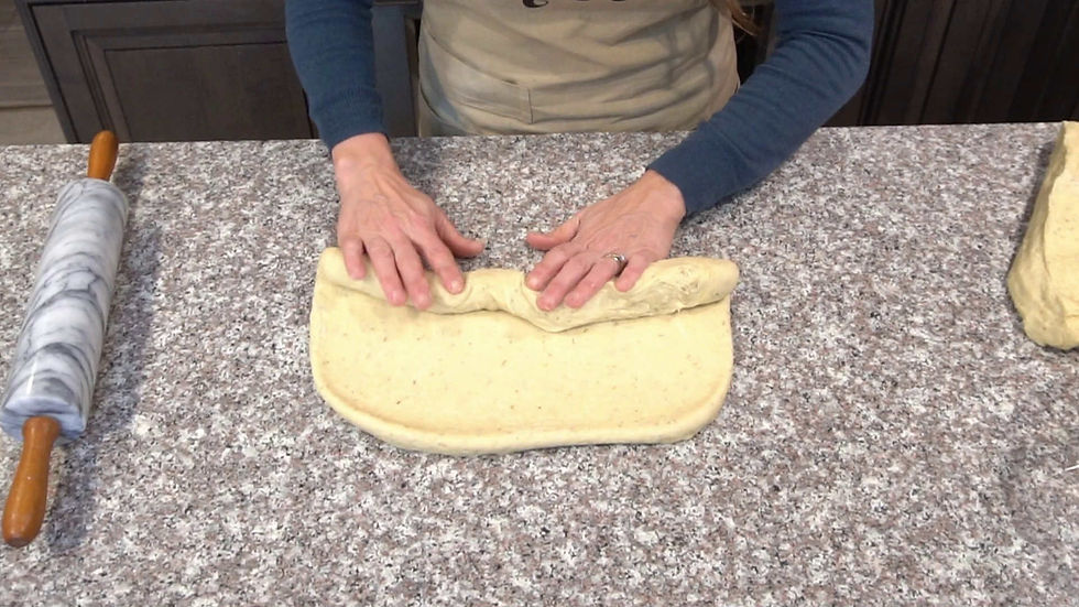 Hands roll dough on a speckled countertop next to a marble rolling pin. Person wears a blue shirt and beige apron, focused on work.