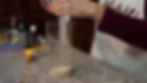 Hands pour ingredients into a tall glass on a granite counter with bottles and a measuring cup. Person wears a maroon sleeve and apron.