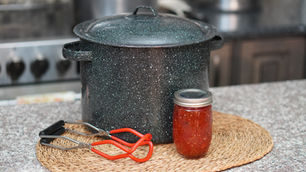 Large speckled pot, red jar tongs, and a sealed jar of red sauce on a woven mat atop a granite countertop in a kitchen setting.