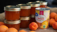 Jars of apricot pineapple jam with gold lids stacked next to a can of crushed pineapple. Apricots scattered around on a burlap cloth.