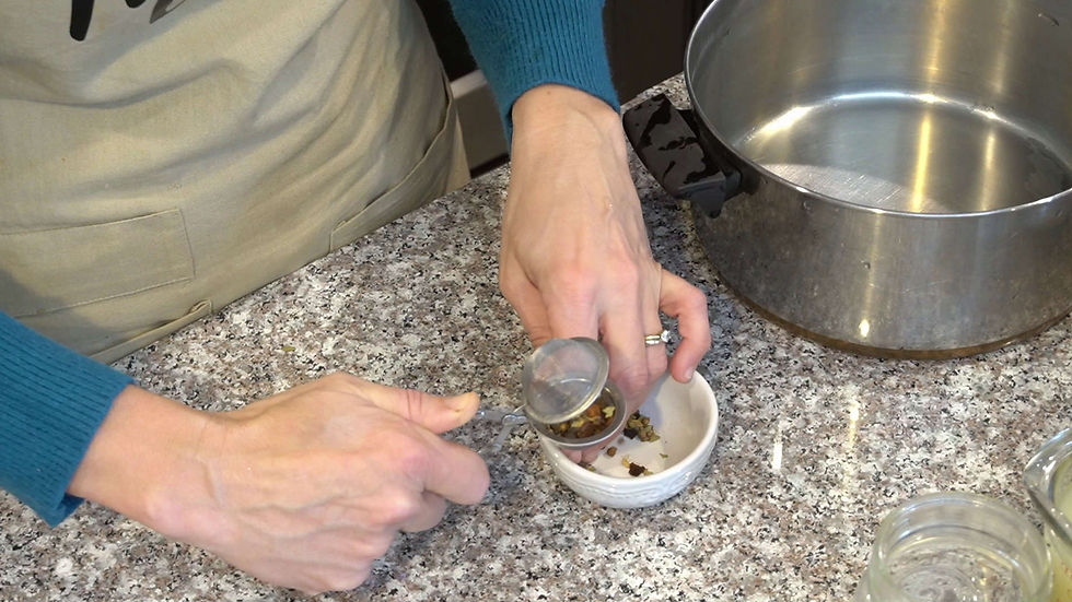Hands filling a tea infuser with herbs over a small white bowl on a granite countertop, next to a metal pot. Blue sleeve and tan apron visible.
