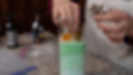 Hands arranging soaps on colored bath salts in a glass jar. Bottles and bags scattered on a granite countertop. Brown and green tones.