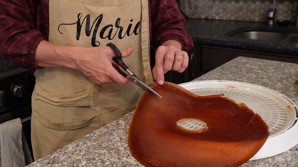 Person in an apron labeled "Marie," cutting dried fruit leather on a granite countertop with scissors. Kitchen setting with focus on hands.