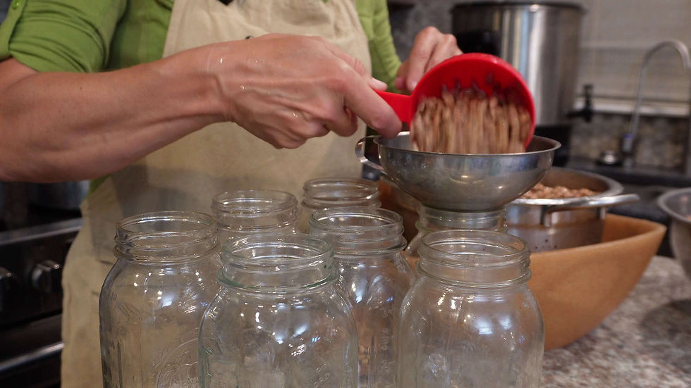 Hands scooping beans into jars with a red measuring cup; kitchen setting, green shirt, and beige apron visible. Mood: focused.