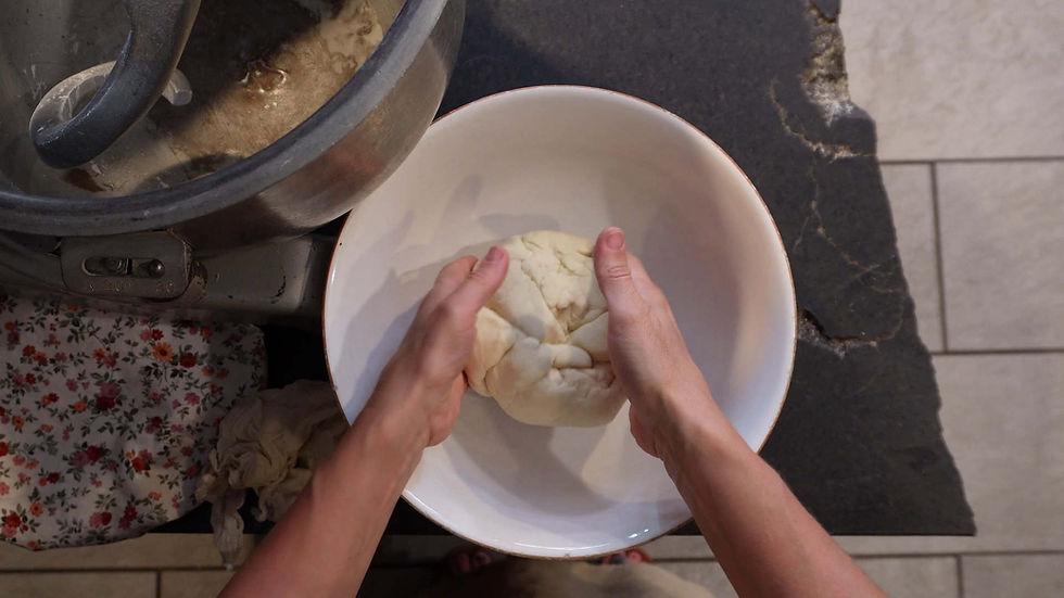 Hands kneading dough in a white bowl on a black table. Nearby, a mixer and floral-patterned cloth add a cozy kitchen vibe.