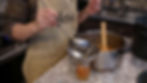 Person in an apron ladling cooked fruit into a jar through a funnel. Kitchen setting with a pot and wooden spoon on a granite countertop.