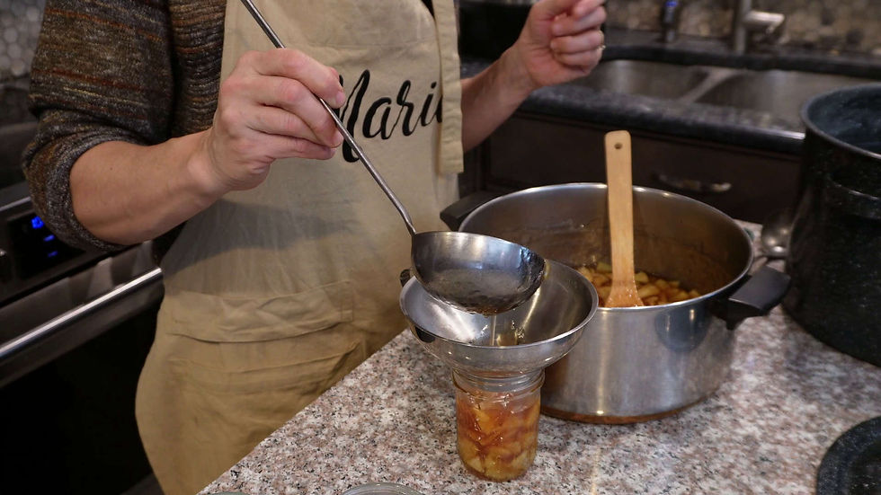 Person in an apron ladling cooked fruit into a jar through a funnel. Kitchen setting with a pot and wooden spoon on a granite countertop.