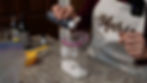 Person in a "Marie" apron pours liquid from a bottle into a zip-top bag of sugar on a speckled countertop, surrounded by kitchen items.