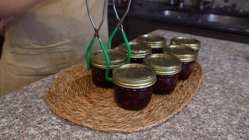 Canning jars of red jam with gold lids sit on a woven mat. Green-lid tongs are held by a person in a kitchen setting.