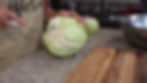 Person cutting a large cabbage on a granite countertop, wearing a beige apron. A wooden cutting board and metal bowl are nearby in the kitchen.