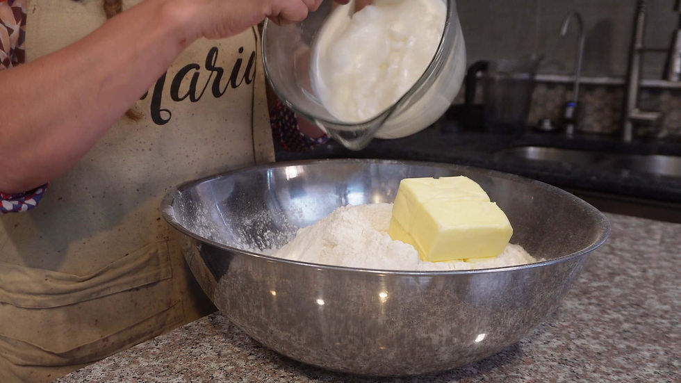 Person in kitchen apron pours cream into a silver bowl with flour and butter on a speckled counter. "Mari" is partly visible on apron.