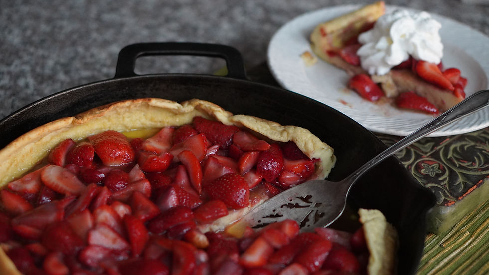 Strawberry-filled Dutch baby pancake in a skillet with a slice served on a plate, topped with whipped cream. Warm and inviting setting.