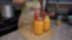 Person in an apron lifts a jar of orange liquid with tongs on a kitchen counter. Stove in background, text reads "Marie." Cozy setting.