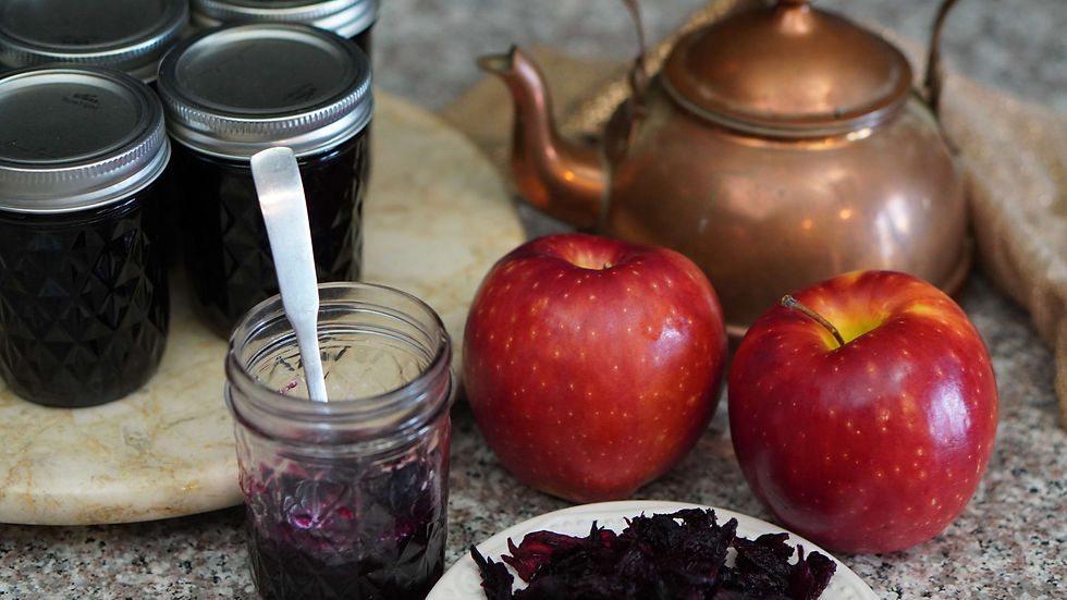 Jars of dark jam with lids, spoon in an open jar, two red apples, and a copper teapot on a marble surface. Plate of dried petals nearby.