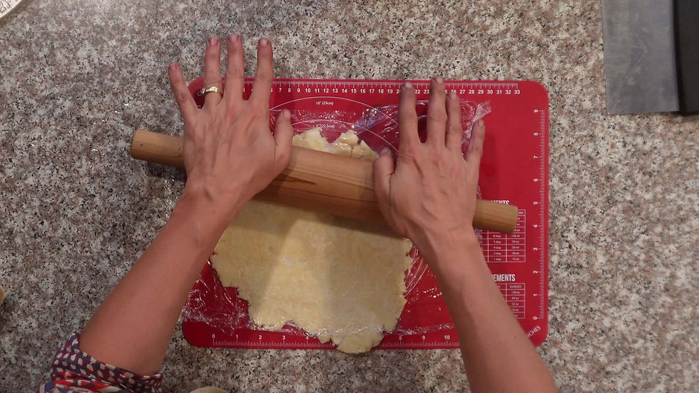 Hands rolling dough on a red mat with measurements, on a speckled countertop. A rolling pin smooths the dough covered in plastic wrap.