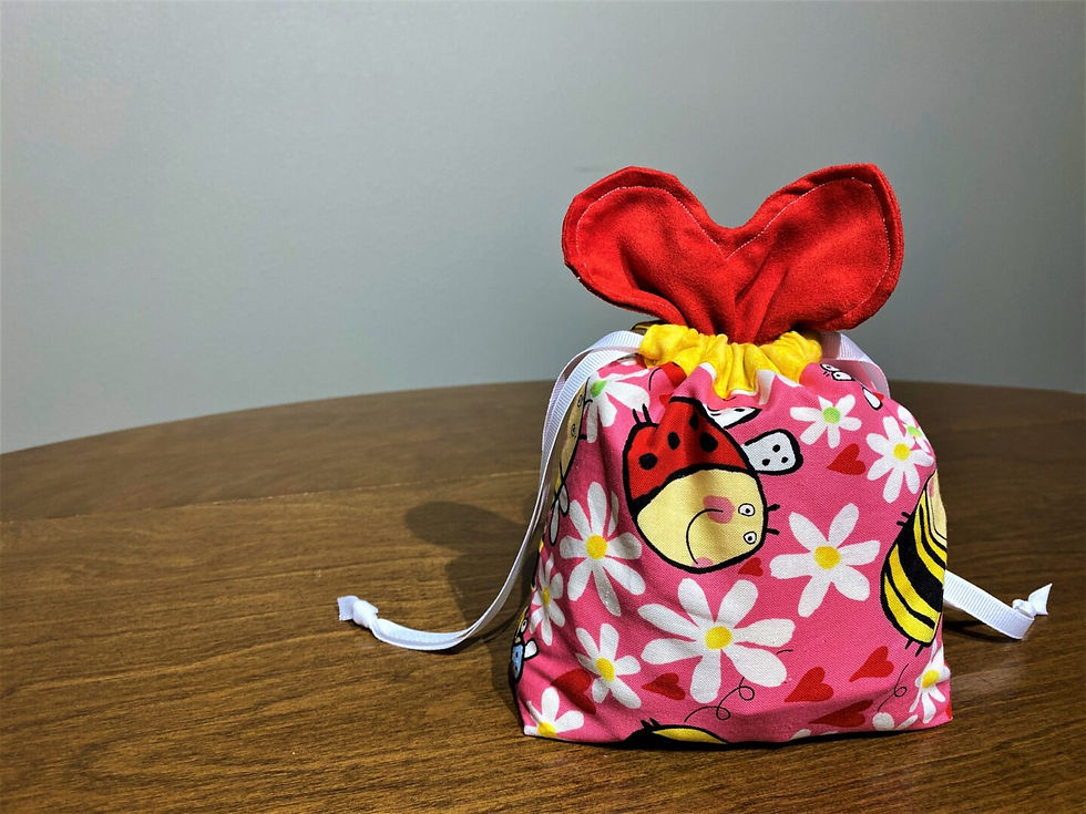 Colorful fabric gift bag with red ears and drawstrings, featuring ladybugs, daisies, and hearts on a pink background, placed on a wooden table.