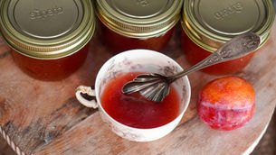 Three jars of red jam with metal lids labeled superb and quebec, a teacup of jam with spoon, and a plum on a tray.