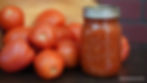 A jar of homemade tomato sauce next to fresh tomatoes on a dark surface. Visible text on jar. Warm, rustic kitchen setting.