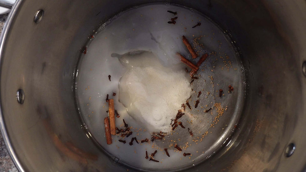 A pot with water, floating mustard seeds, cinnamon sticks, and cloves. Sugar is dissolving, creating a cooking mixture.