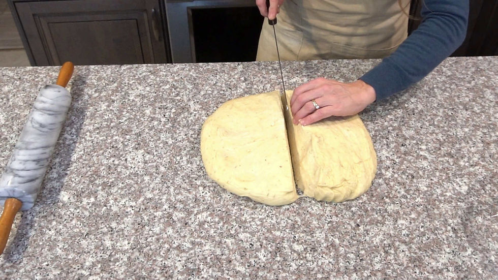Hands slicing dough on a granite countertop with a knife, next to a marble rolling pin. Gray and neutral tones, indoor kitchen setting.