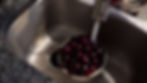 Cherries being rinsed under running water in a silver sink. The sink and faucet are metal, and the countertop is dark speckled granite.