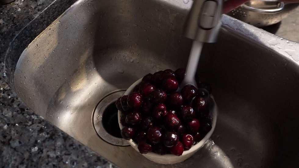 Cherries being rinsed under running water in a silver sink. The sink and faucet are metal, and the countertop is dark speckled granite.