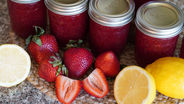 Jars of red jam sit on a woven mat surrounded by fresh strawberries and lemon halves. The lids read "superb." Bright and vibrant scene.
