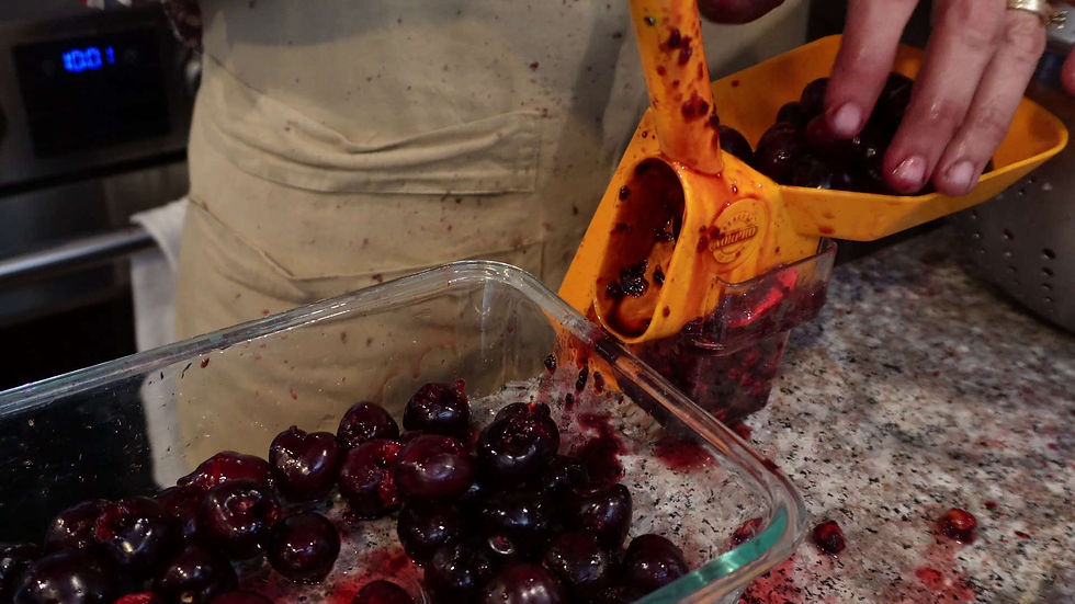 Hands use an orange cherry pitter on a countertop, spilling cherry juice and pits into a glass dish. A beige apron is in the background.