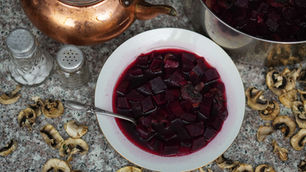 Bowl of red beet soup with a spoon, surrounded by dried mushrooms, salt and pepper shakers, and a copper kettle on a speckled countertop.