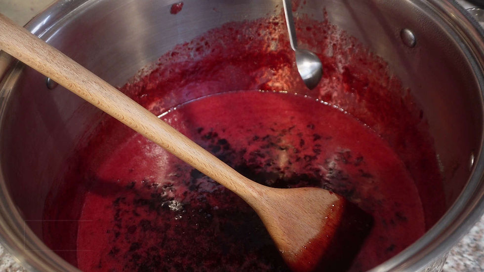 Wooden spoon stirring deep red sauce in a stainless steel pot. A silver spoon rests inside. Bubbly texture suggests cooking.
