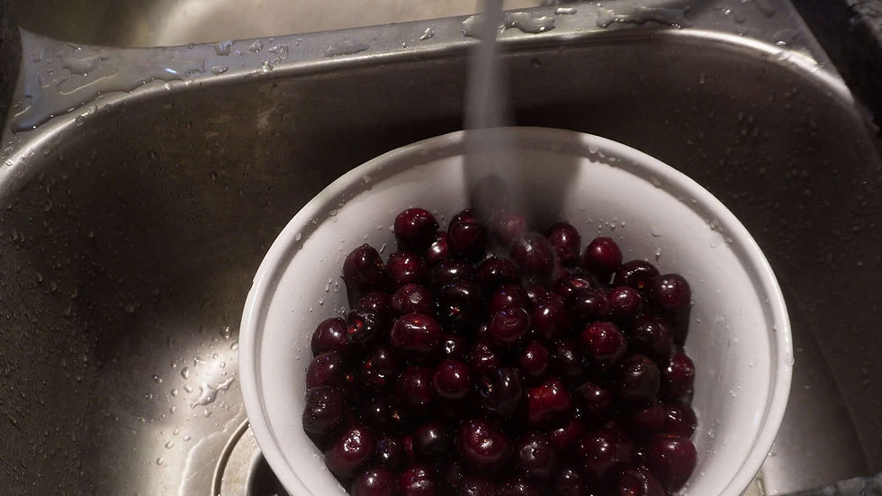 Bowl of dark cherries being rinsed under running water in a stainless steel sink. Water droplets visible, creating a fresh, clean mood.