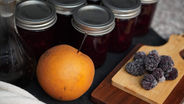 Jars of red jam with metal lids, an orange pear, and frozen blackberries on a wooden board. A dark liquid in a glass bottle nearby.