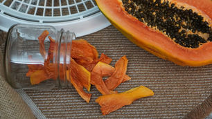 Sliced papaya in a jar spills onto a brown fabric. A halved papaya with black seeds is nearby, set on a woven textured background.