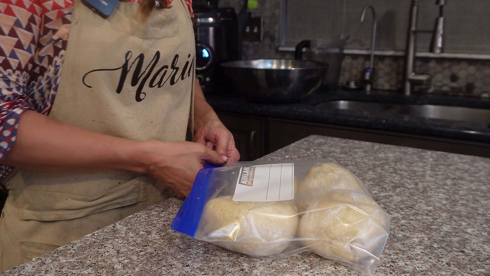 Person in a patterned shirt seals dough in a labeled plastic bag on a granite kitchen counter. Background has a sink and a bowl.