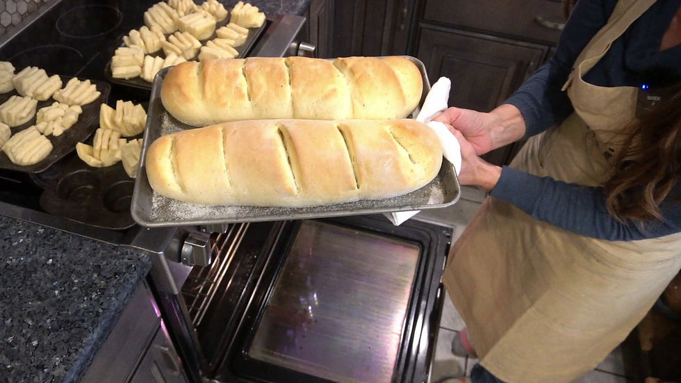 Person in apron holds a tray with two freshly baked golden loaves. Background shows an open oven and trays of uncooked dough in a kitchen.