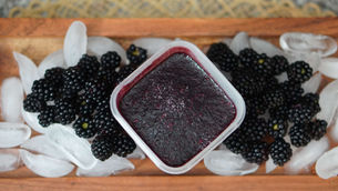 A container of blackberry freezer jamis surrounded by fresh blackberries and ice on a wooden tray. The setting is cozy and rustic.