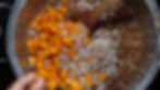 Chopped apricots and sugared nuts in a pot with syrup, stirred with a wooden spoon. A hand is visible, suggesting cooking action.