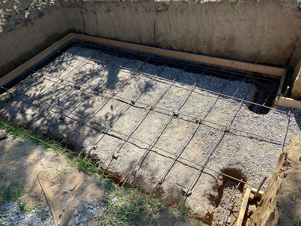 Excavated foundation pit with steel rebar grid and wooden forms over gravel, ready for a concrete pour in bright sunlight