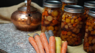 Jars of canned carrots on a granite counter with fresh carrots and a copper kettle. Warm, rustic kitchen setting.