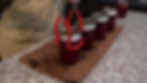 Person in apron uses red tongs to handle jar of homemade jam on a textured counter. Four jars visible, creating a cozy kitchen scene.