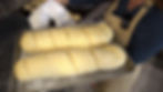 Two unbaked baguettes on a tray held by a person in an apron, kitchen setting. Dough is scored, counter with dough pieces visible.