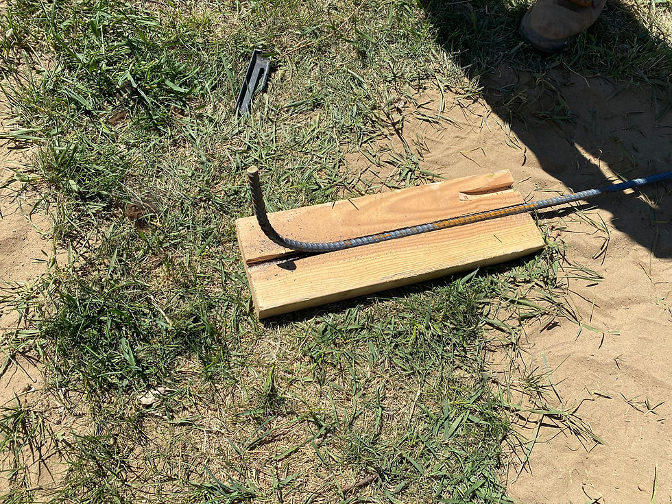 Bent rebar laid across a wooden block on patchy grass and dirt, with a boot shadow nearby in bright sunlight.