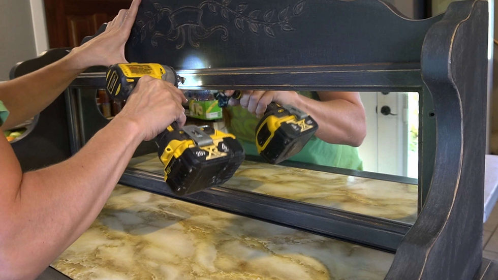 Hands use two yellow cordless drills to assemble a dark wooden dresser with mirror in a home interior.