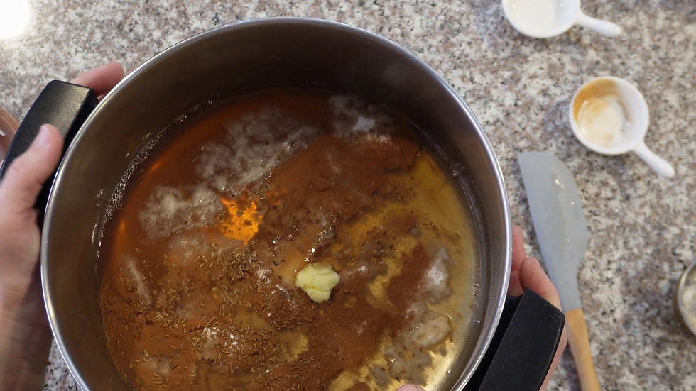 Hands holding a pot with melted butter and spices on a speckled countertop. Empty cups and a spatula are visible. Warm, cooking atmosphere.