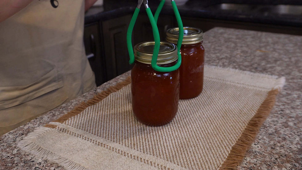 A person uses tongs to lift a jar of red sauce on a fringed beige cloth. Another jar is next to it on a granite countertop.
