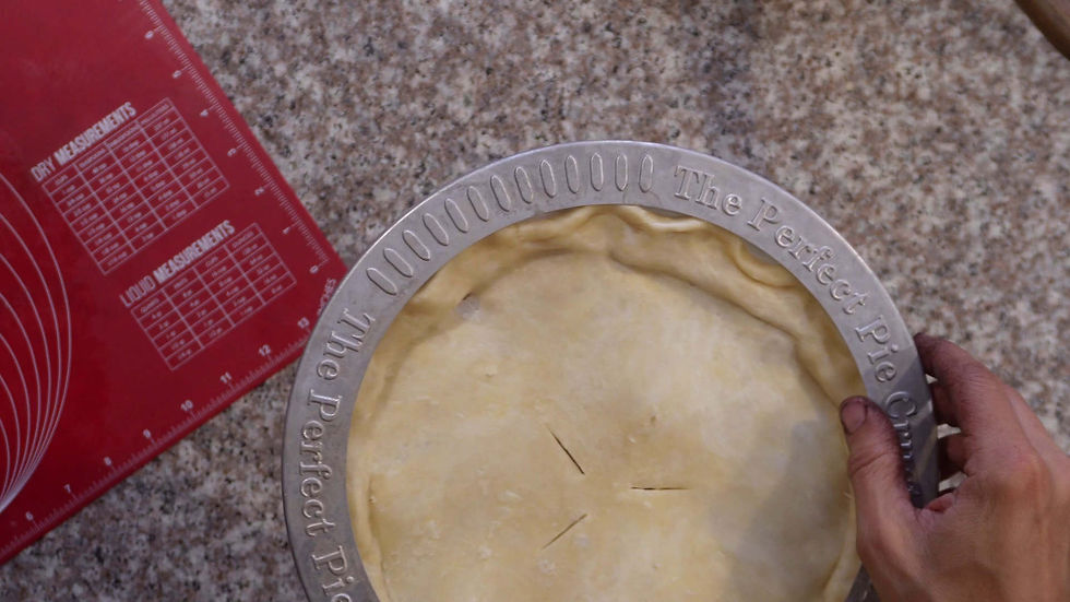 Hands hold a pie crust in a pan labeled "The Perfect Pie Crust" on a granite countertop. A red measuring mat is partially visible.