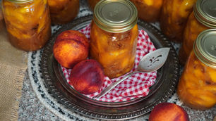 Jars of canned peaches on a red-checkered cloth with fresh nectarines and a decorative spoon on a silver tray, set on a woven mat.