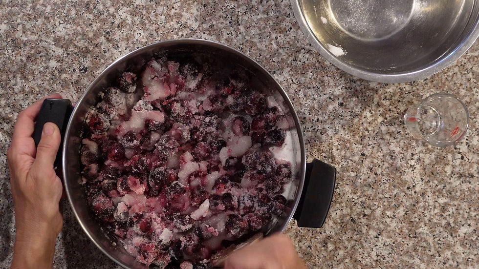 Hands stirring sugar-coated cherries in a metal pot on a speckled countertop. A metal bowl and measuring cup are nearby.