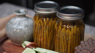 Two jars of pickled asparagus beside a gray vase on a wooden tray. A green patterned cloth and brown object are in the foreground.